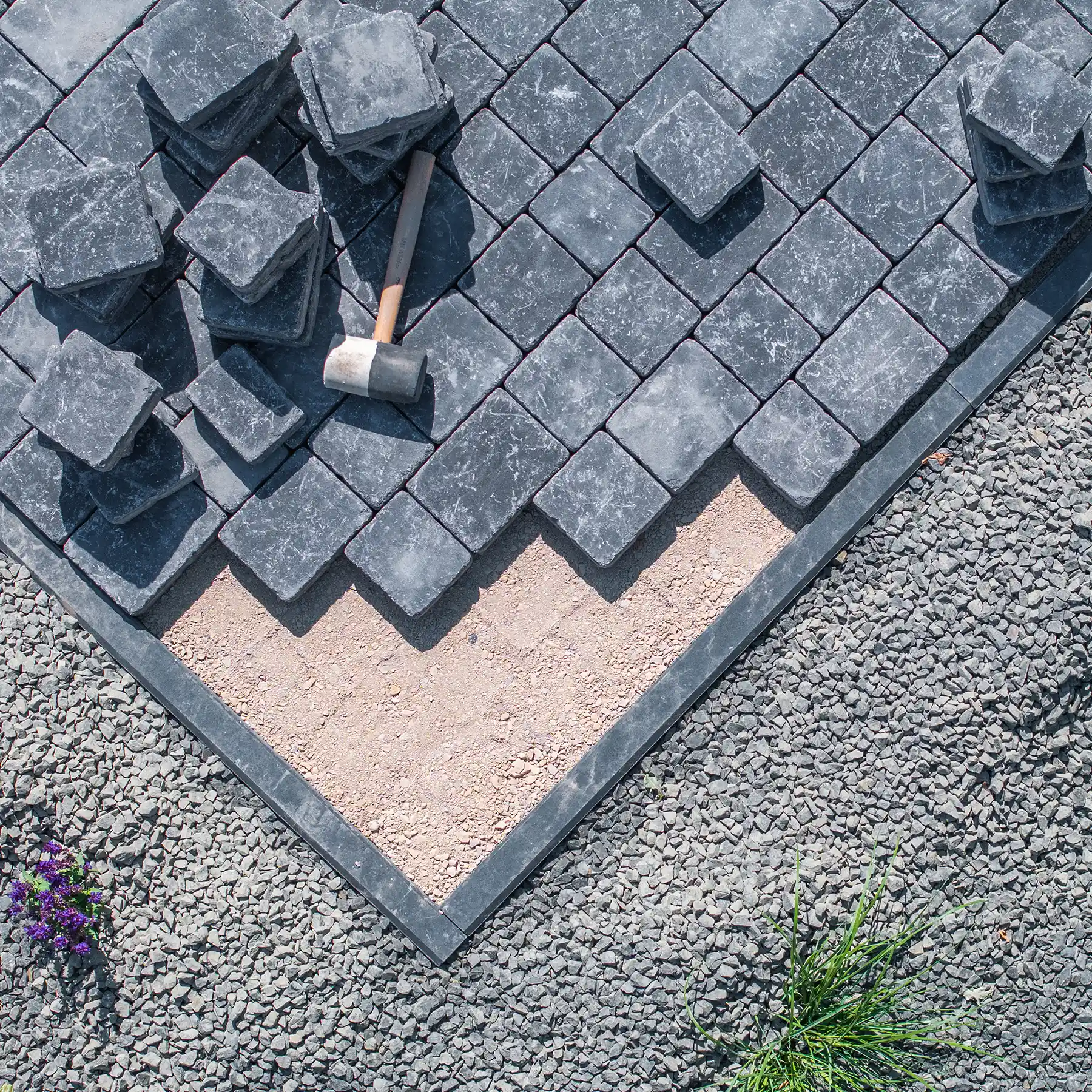 Pflasterarbeiten mit dunklen Natursteinplatten, Verlegehammer, Splittbett und Kiesfläche.