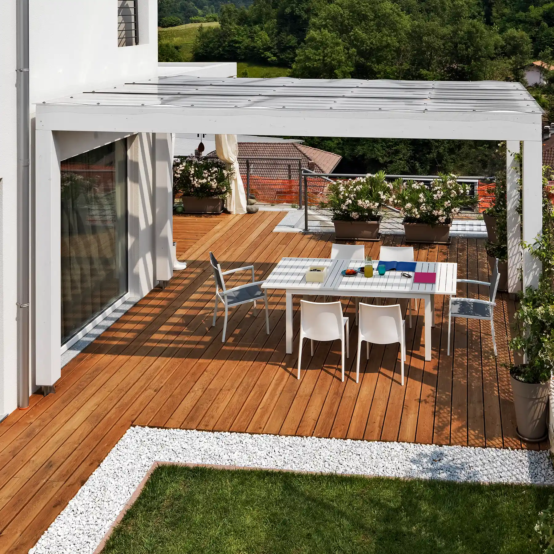 Moderne Holzterrasse mit Überdachung, Esstisch, Pflanzkübeln, Rasenfläche und weißem Kiesrand.
