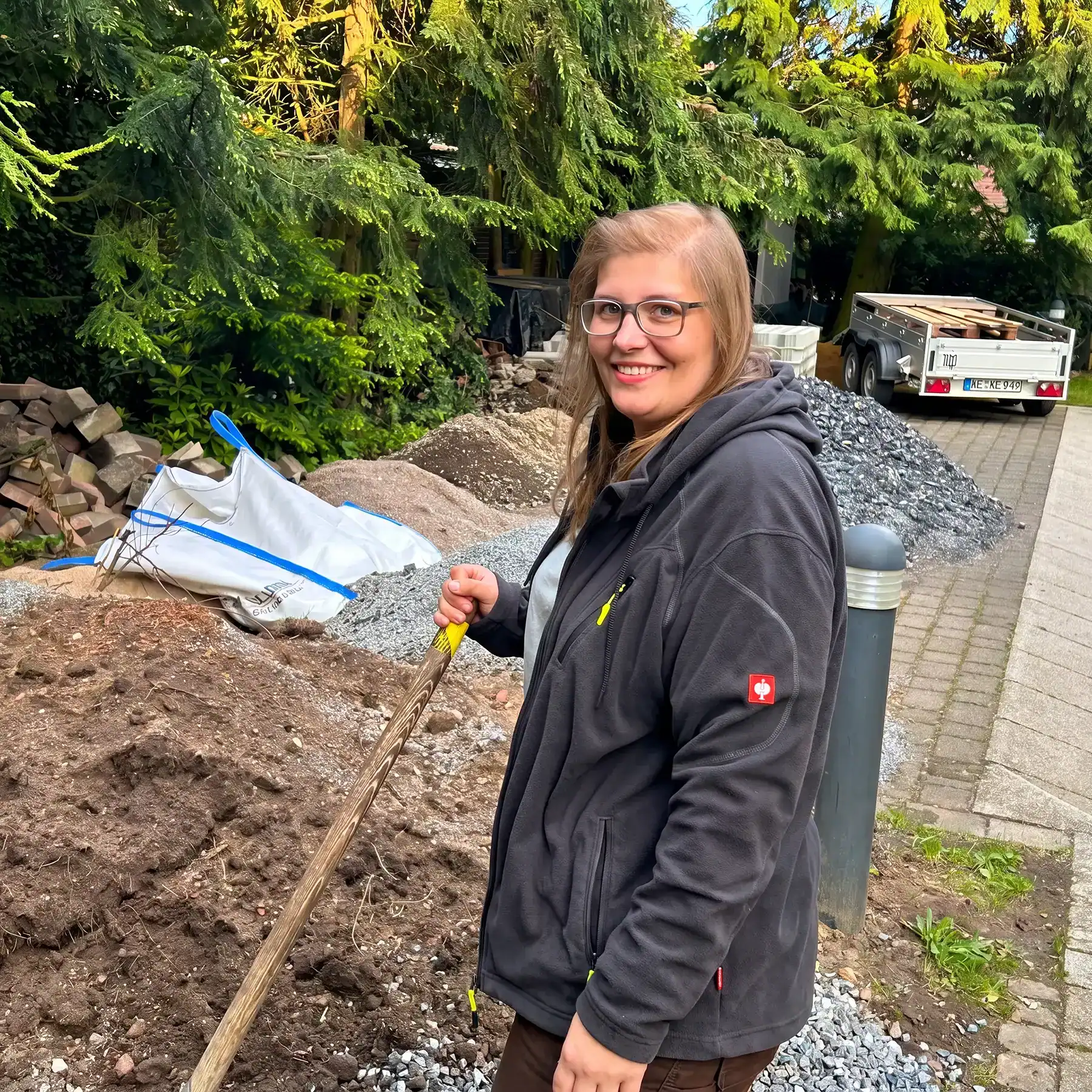 Person mit Arbeitskleidung und Werkzeug bei Erdarbeiten auf einer Baustelle im Grünen.